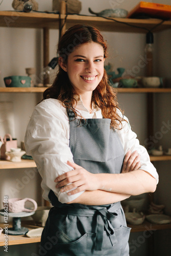 Happy young potter stands and smiling with shelves filled with earthenware in the background in her pottery studio. Craftswoman running a successful small business.