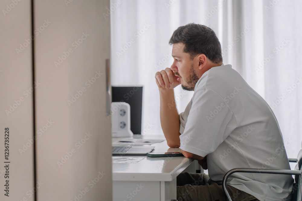 A man in a white shirt working at a desk, deeply focused, with a laptop and smartphone in a bright, minimalistic indoor setting. A man in a white shirt working at a desk, deeply focused, with a laptop and smartphone in a bright, minimalistic indoor setting.