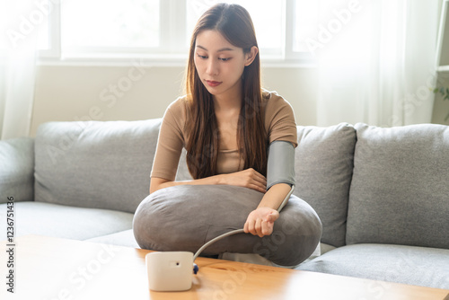 Hypotension, asian young woman sitting on couch checking blood high pressure and heart rate with digital pressure monitor machine making self check up on her arm at home. Healthcare and medical. photo