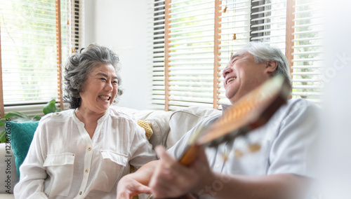 Fotografie Senior Asian couple laughing together while sitting on a sofa, enjoying music with a guitar in a cozy living room with sunlight streaming through blinds, reflecting happiness and companionship