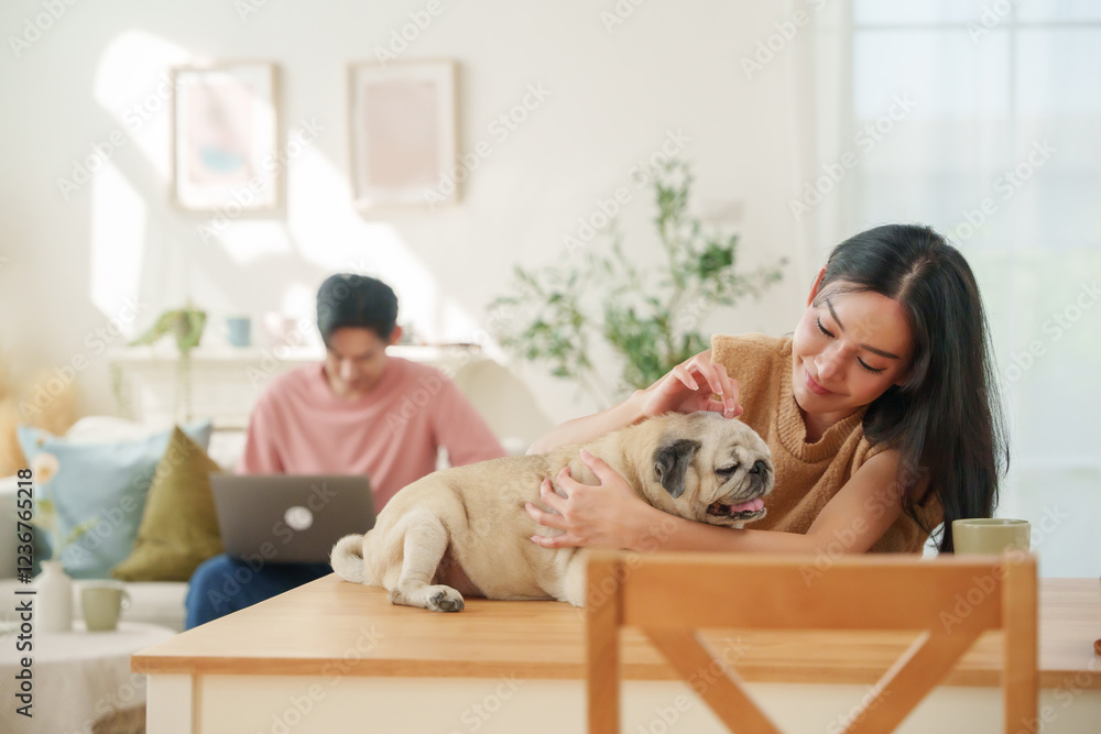 Young Asian couple spending a relaxed morning with their pug dog at home, woman gently cuddling the dog at the kitchen table while the man works on his laptop in the bright living room. Young Asian couple spending a relaxed morning with their pug dog at home, woman gently cuddling the dog at the kitchen table while the man works on his laptop in the bright living room.