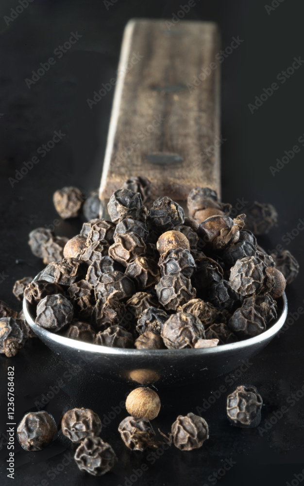 Raw organic black peppercorns or Dry black pepper seeds in stainless steel spoon on black background. use it as your Space for text, Design, Wallpaper and Poster, Selective focus.