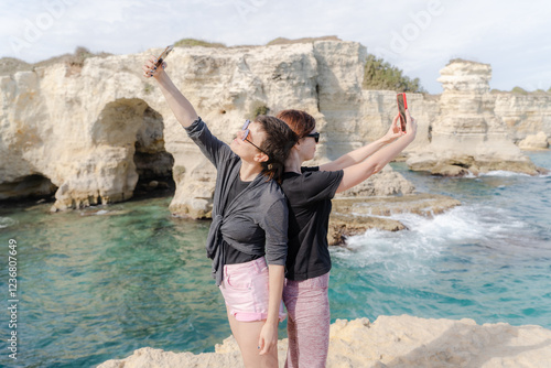 Two girls stand with their backs to each other against the backdrop of the sea and film themselves on their phones.