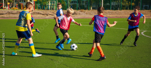 The photo shows a group of young soccer players in colorful jerseys on a green turf field. They are actively playing and competing for the ball, with some players in motion and others watching.