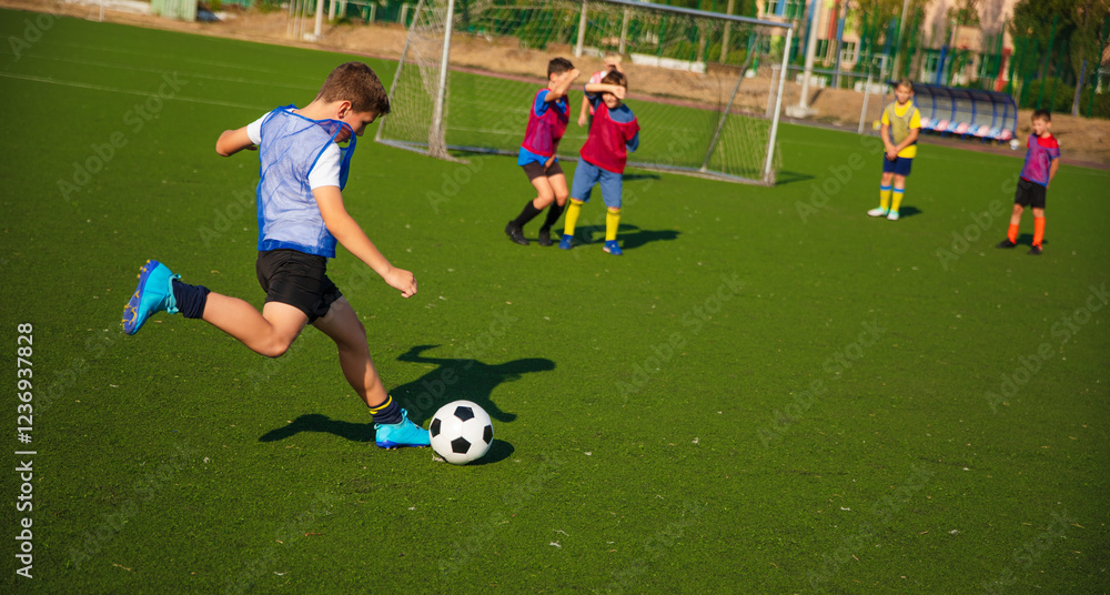 Obraz premium The photo shows a young soccer player in action, kicking a black-and-white ball on a green turf field. Other players in colorful jerseys are positioned in the background, ready for the next play.