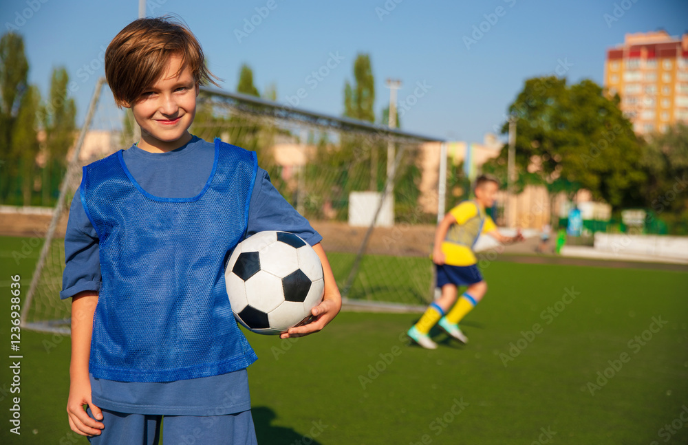 A young soccer player in a blue training vest holds a soccer ball, smiling at the camera. In the background, another player in a yellow shirt is in motion, with a goalpost and clear skies visible. A young soccer player in a blue training vest holds a soccer ball, smiling at the camera. In the background, another player in a yellow shirt is in motion, with a goalpost and clear skies visible.