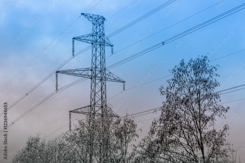 Fototapeta premium A high-voltage electricity pylon with power lines stretching across the sky during dusk. The silhouette contrasts with the gradient hues of blue and orange in the background.