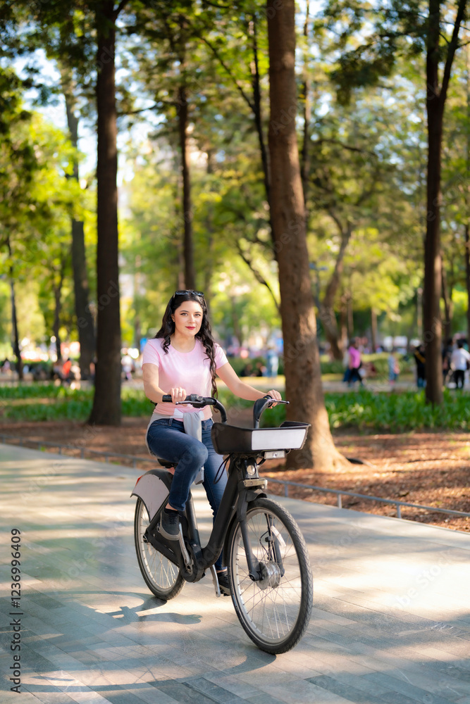 Happy young Latina woman with wavy hair, pink t-shirt, and jeans rides an Ecobici through Alameda Central, Mexico City, enjoying a sunny day surrounded by nature and people. Happy young Latina woman with wavy hair, pink t-shirt, and jeans rides an Ecobici through Alameda Central, Mexico City, enjoying a sunny day surrounded by nature and people.