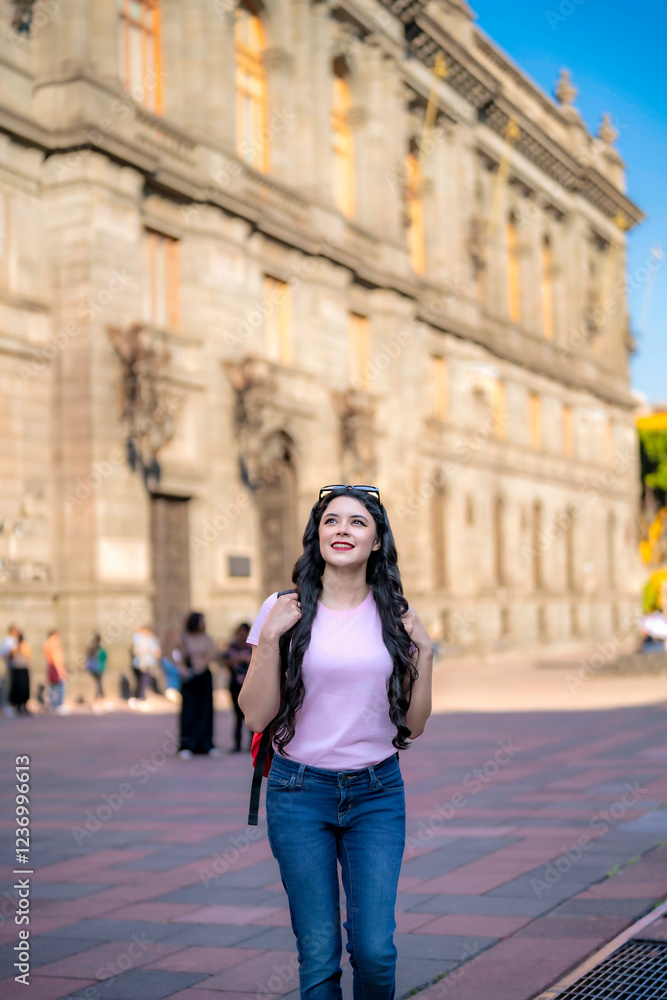 Fototapeta premium Happy young Latina woman with wavy hair, pink t-shirt, jeans, and a red backpack walks in front of the MUNAL museum in Mexico City, enjoying a sunny day and historic architecture.