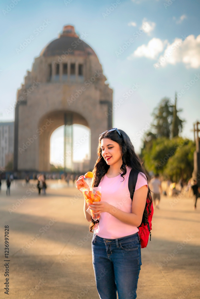 Fototapeta premium Smiling young Latina woman with wavy hair, pink t-shirt, jeans, and a red backpack enjoys spicy mango in front of the Monument to the Revolution in Mexico City during a sunny afternoon. 