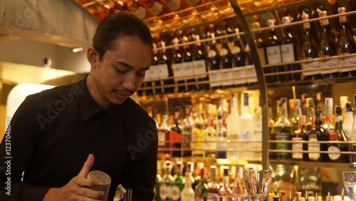 Smiling Asian smart bartender shaking with two hands in mixed cocktail in shaker preparing at luxurious counter bar, preparation special refreshment alcohol drink nightlife warm nightclub. Vinosity. photo