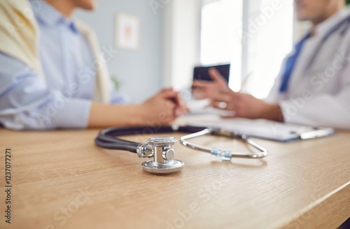 Obraz na plátně Close up shot of a stethoscope on a wooden table, with a doctor and patient talking in a blurred background