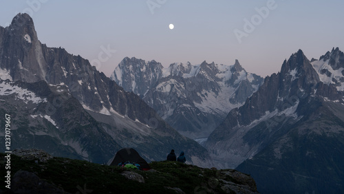 A couple watches the sunset in the mountains with the moon in the sky