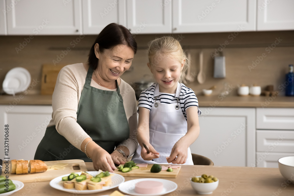 Loving granny teach to cook little grandkid girl, wear aprons stand at dining table cutting fresh cucumbers, preparing delicious sandwiches for breakfast. Older gen teaching to younger, share skills Loving granny teach to cook little grandkid girl, wear aprons stand at dining table cutting fresh cucumbers, preparing delicious sandwiches for breakfast. Older gen teaching to younger, share skills