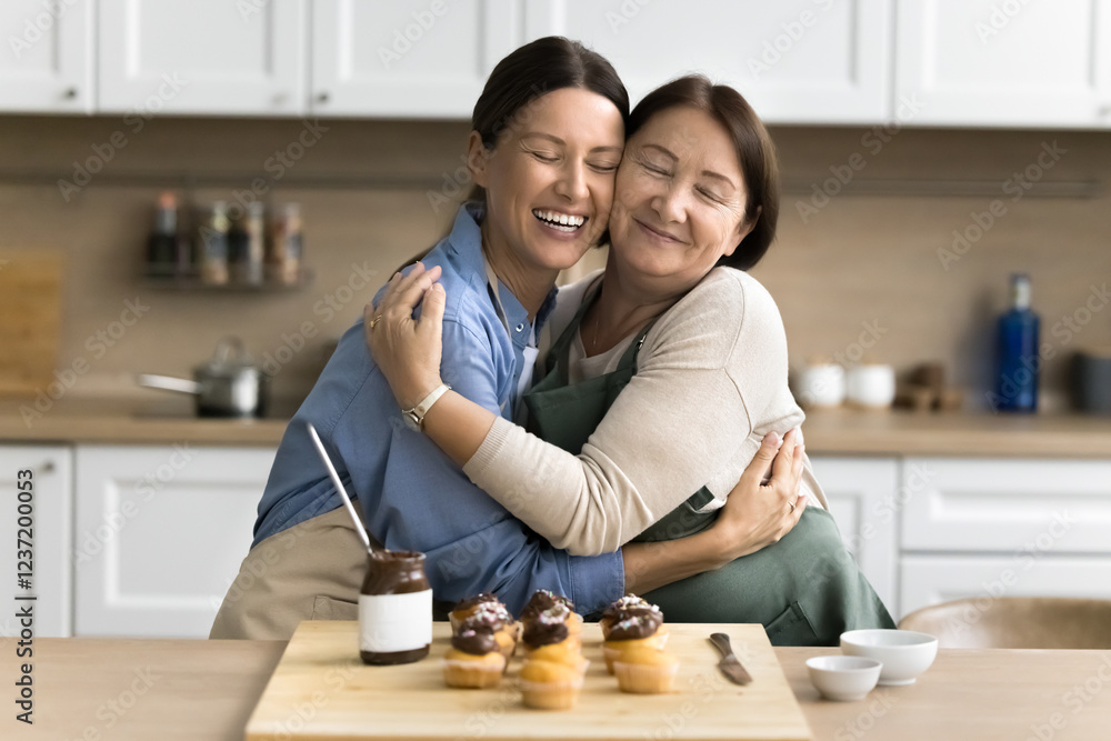 Fototapeta premium Young adult daughter cuddling tightly her older smiling mother, women wear aprons standing together, hugging, while cooking together sweet chocolate muffins, family recipe dessert on weekend at home
