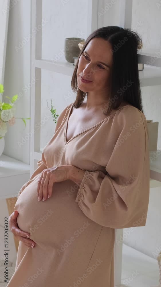 Cheerful pregnant middle aged Caucasian woman smooths a baby bump and dreaming at home. Motherhood planning concept. Vertical video.