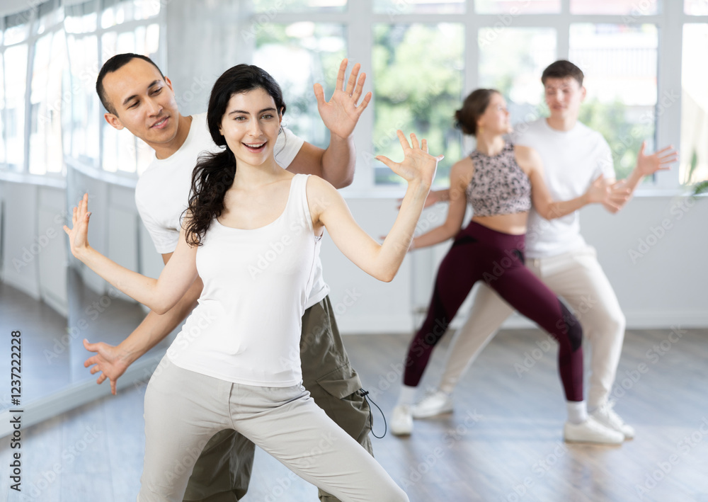 Naklejka premium Cheerful young couple in casual workout attire enjoying dancing during group training in dance studio, practicing playful Charleston. Active hobbies concept