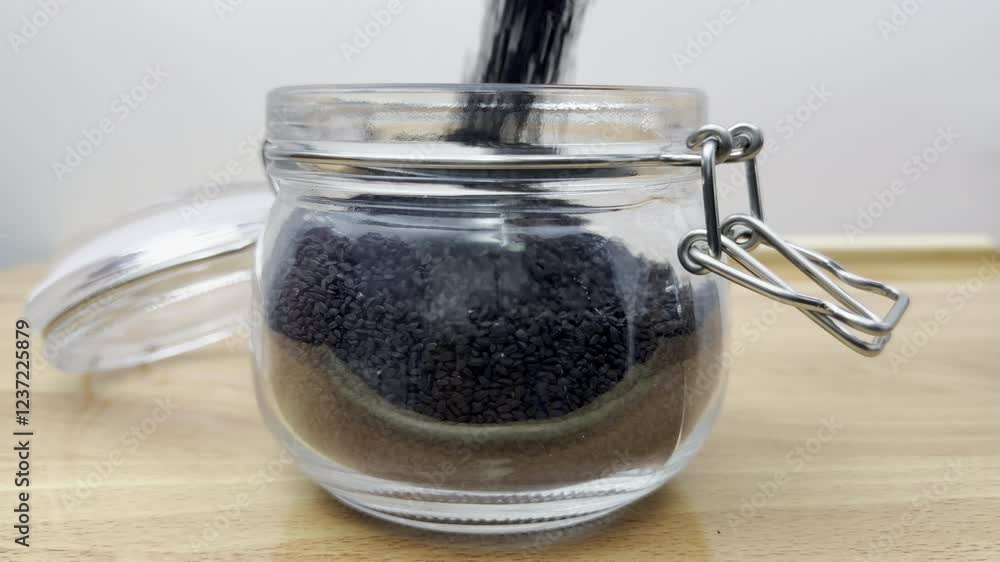 Closeup slow motion shot of tiny, black nigella seeds (also kalonji and kalwanji)) pouring into a glass storage jar. Generally used as an ingredient or spice or seasoning for cooking. Closeup slow motion shot of tiny, black nigella seeds (also kalonji and kalwanji)) pouring into a glass storage jar. Generally used as an ingredient or spice or seasoning for cooking.