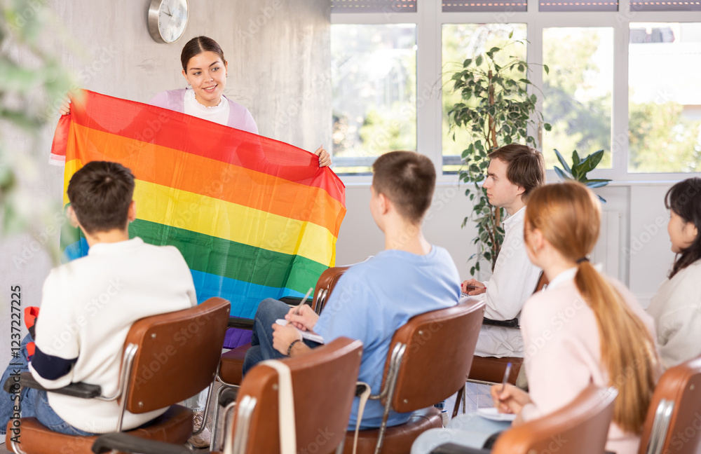 Obraz premium Smiling young female student talking about international LGBT social movements during lecture in auditorium, showing colorful rainbow flag