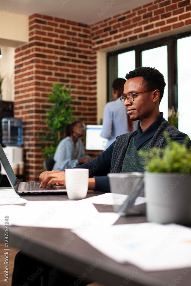 © DC Studio - Black businessman seated at desk, using his digital device to reply to work emails and review daily tasks. Male entrepreneur typing on his laptop, preparing startup business project in modern office.