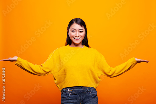 Beautiful woman smiles happily, using hand gestures to compare two options, against yellow background. Asian lady cheerfully demonstrating different objects on open palms, presenting and advertising.