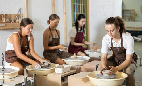 Concentrated young guy in apron working with pottery wheel in workshop with other persons engaged in pottery photo