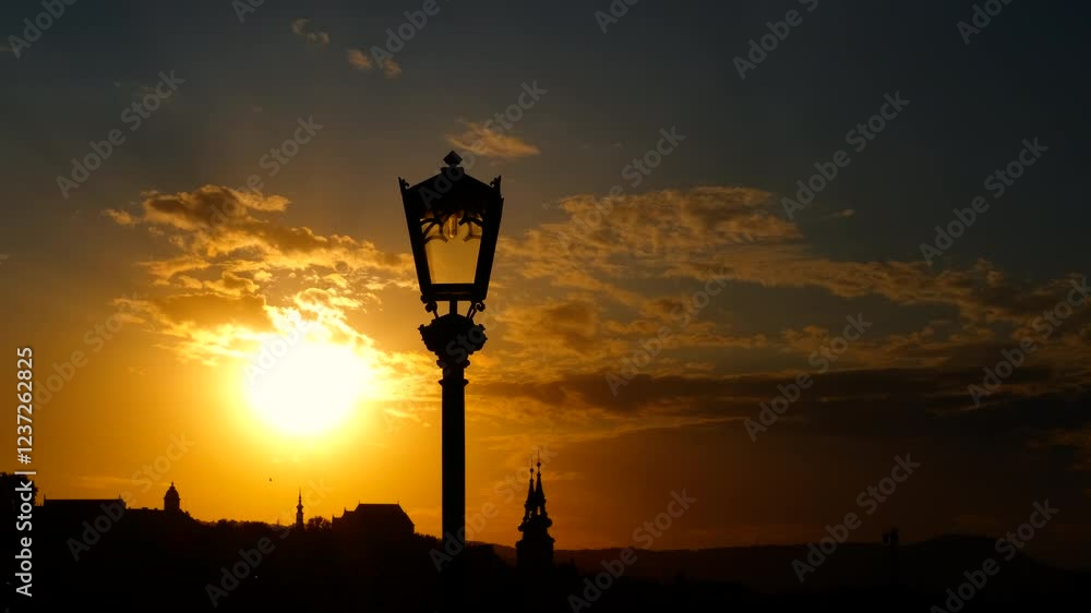 The sun is setting behind a silhouetted cityscape with a streetlamp in the foreground. The sky is a beautiful orange and yellow The sun is setting behind a silhouetted cityscape with a streetlamp in the foreground. The sky is a beautiful orange and yellow