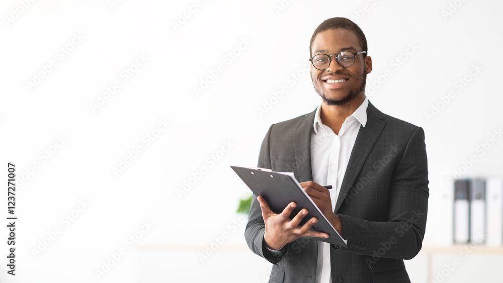 Happy young black male psychologist writing in clipboard, looking at camera and smiling in modern office. Psychotherapy services, mental health professional concept Happy young black male psychologist writing in clipboard, looking at camera and smiling in modern office. Psychotherapy services, mental health professional concept