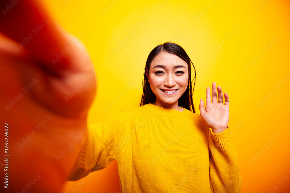 Smiling asian lady making a hello gesture towards the camera, vlogging for her online audience. Beautiful woman waving her hand and taking a selfie against yellow background, pov concept. Smiling asian lady making a hello gesture towards the camera, vlogging for her online audience. Beautiful woman waving her hand and taking a selfie against yellow background, pov concept.