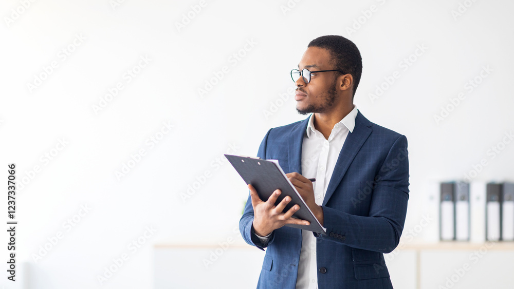 Serious young black psychologist writing in clipboard, looking aside deep in thought at modern office. Psychotherapy services, mental health professional concept Serious young black psychologist writing in clipboard, looking aside deep in thought at modern office. Psychotherapy services, mental health professional concept