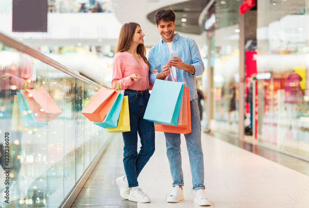 Cheerful Spouses Shopping Using Mobile Phone Posing Holding Colorful Shopper Bags In Modern Mall Indoors. Buyers Couple Purchasing Clothes Online Via Application. Sales Concept. Full Length Cheerful Spouses Shopping Using Mobile Phone Posing Holding Colorful Shopper Bags In Modern Mall Indoors. Buyers Couple Purchasing Clothes Online Via Application. Sales Concept. Full Length