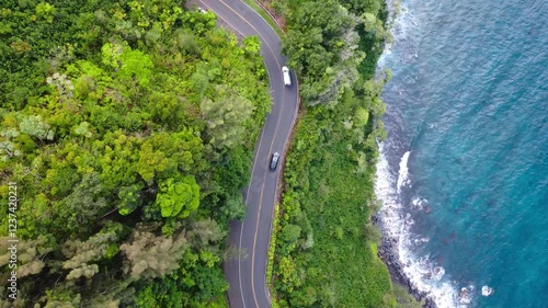 An aerial video of cars driving down a winding road along a lush tropical coastline with crystal clear turquoise water. Road to Hana, Maui Hawaii. 