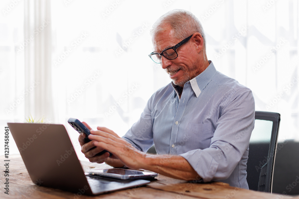 Handsome smiling senior man wearing glasses using mobile phone while sitting at his cozy workplace with laptop at office, retired male chatting with friends in social media, typing on smartphone Handsome smiling senior man wearing glasses using mobile phone while sitting at his cozy workplace with laptop at office, retired male chatting with friends in social media, typing on smartphone