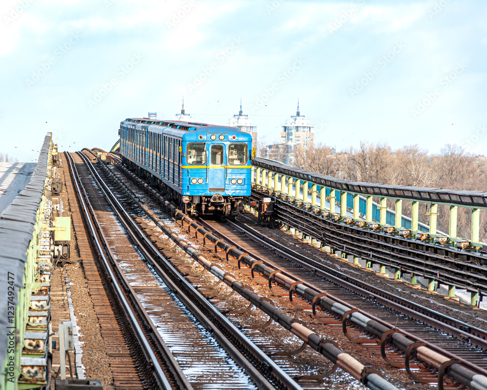 Obraz premium A vibrant blue Kyiv metro train traveling across a bridge, framed by snowy tracks and city architecture in the distance.