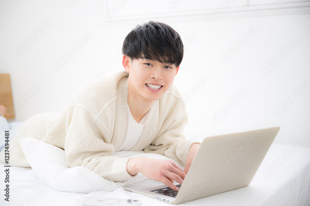 ベッドの上でうつ伏せで寝転がりパソコンをする笑顔の20代の若い男性カメラ目線 学生やフリーランスの若手のイメージ ベッドの上でうつ伏せで寝転がりパソコンをする笑顔の20代の若い男性カメラ目線 学生やフリーランスの若手のイメージ