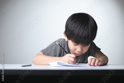 A focused Thai boy writing in a notebook with a pencil, concentrating on his studies at a desk. photo