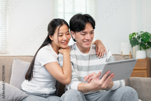 Tablou pe pânză A lovely Asian couple sits together on a sofa in the living room, watching a video on a tablet
