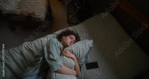 A woman is crying and hugging a pillow lying in bed at home. The mascara is smeared under the eyes. The mobile phone is on her bed