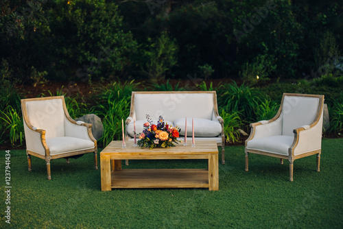 This charming outdoor area features a stylish sofa, comfy armchairs, and a floral centerpiece on a wooden table, all surrounded by lush greenery, creating a serene space for relaxation and gatherings