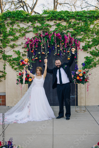 Amidst a beautiful and vibrant floral backdrop, a delighted couple joyfully celebrates their union, marking the special occasion of their wedding with family and friends gathered around them