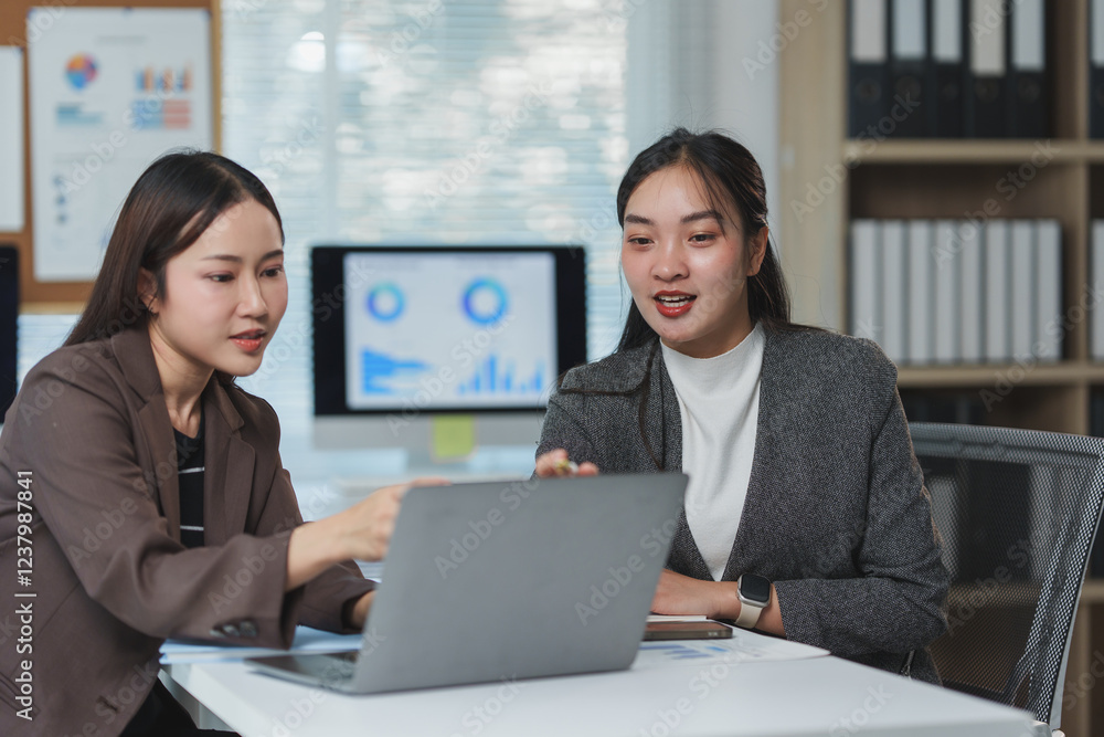 Two professional women engaged in a collaborative work session, discussing data on a laptop in a modern office environment. Charts and graphs visible on a screen in the background Two professional women engaged in a collaborative work session, discussing data on a laptop in a modern office environment. Charts and graphs visible on a screen in the background