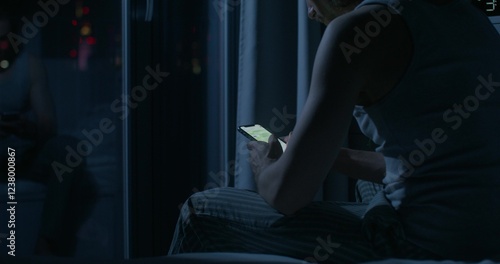 A man uses a mobile phone while sitting on the bed at night. Close-up of his hands, an unrecognizable person. Dark room, home interior