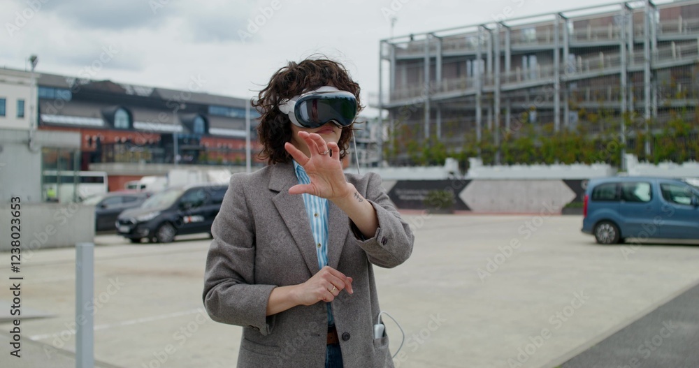 Fototapeta premium A stylish woman in a jacket wearing a virtual reality headset stands on a city street and moves her arms. Summer day