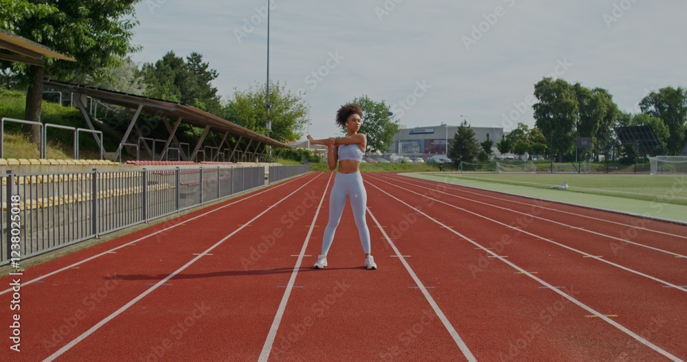 Fototapeta premium A woman in sports clothes and with earbuds in her ears does exercises standing at the city stadium on a summer day