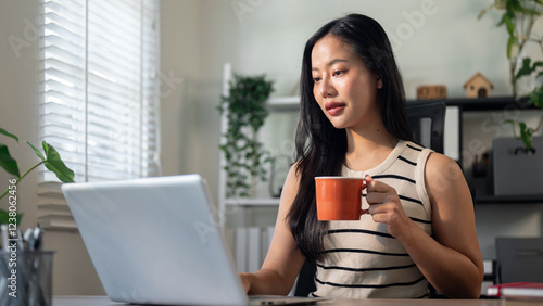 A confident Asian businesswoman is enjoying her morning coffee while working on her laptop.