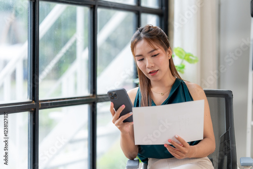 Focused Professional Woman  A young  Asian businesswoman sits by a large window  attentively reviewing documents on a sheet of paper while simultaneously checking her smartphone.
