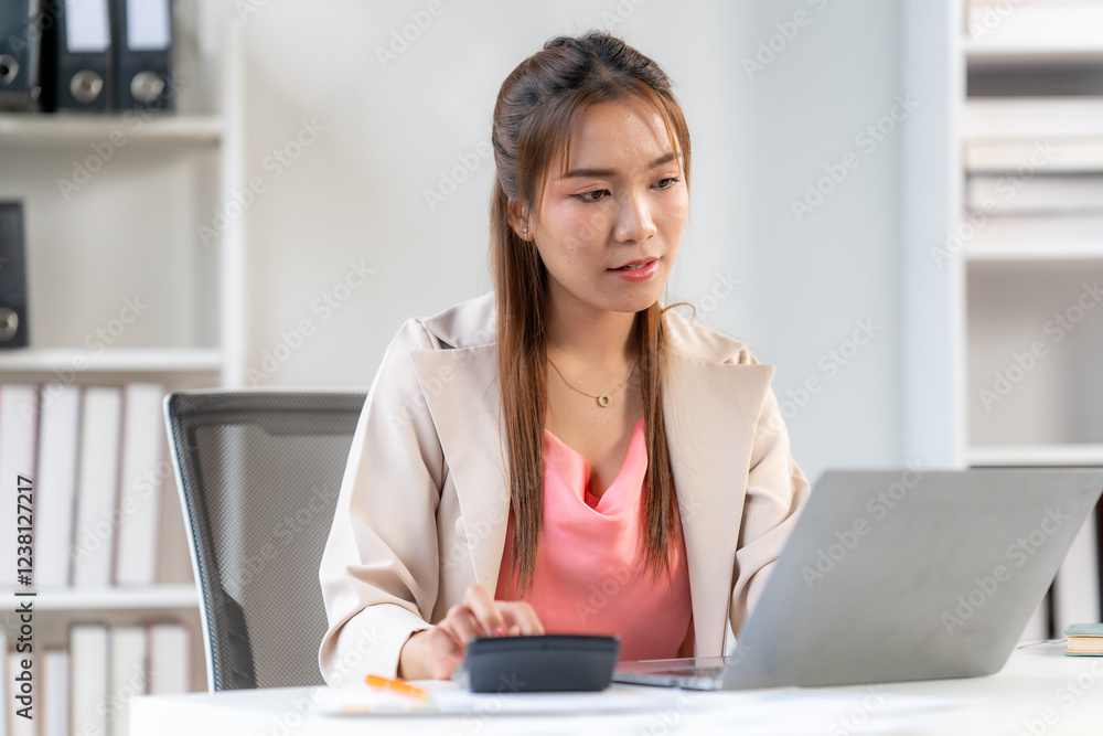 Focused on the Task: A young woman meticulously calculates figures on a calculator, her brow furrowed in concentration as she works on a laptop, showcasing a sense of dedication and focus. Focused on the Task: A young woman meticulously calculates figures on a calculator, her brow furrowed in concentration as she works on a laptop, showcasing a sense of dedication and focus.