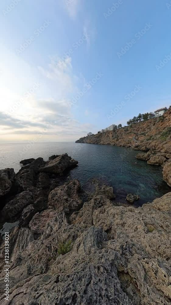 Vertical video. A Male and Female Traveler Sitting on Jagged Rocks Along a Cliffside, Overlooking the Calm Blue Ocean as They Admire the Sunset Behind a Clouded Sky. Peaceful Coastal Moment Vertical video. A Male and Female Traveler Sitting on Jagged Rocks Along a Cliffside, Overlooking the Calm Blue Ocean as They Admire the Sunset Behind a Clouded Sky. Peaceful Coastal Moment