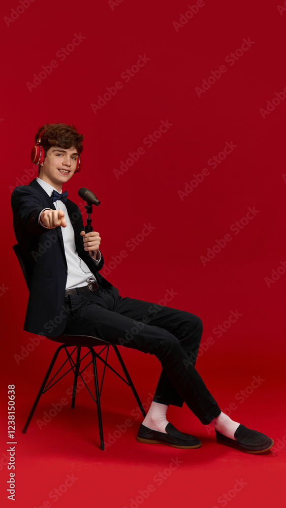 Obraz premium Smiling teen boy, student in black suit and red headphones, sitting on chair with microphone, recording online podcast against red studio background. Concept of youth, education, emotions