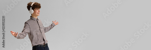 Emotional young boy, teen in grey shirt, wearing headphones and spreading hands in joy and excitement against grey background. Concept of human emotions, youth, lifestyle. Banner. Copy space for ad photo